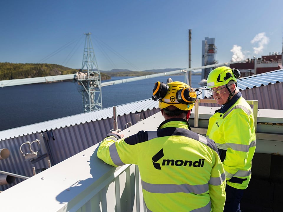 two men in hi-viz clothing overlook a production site with a river and forest in the background