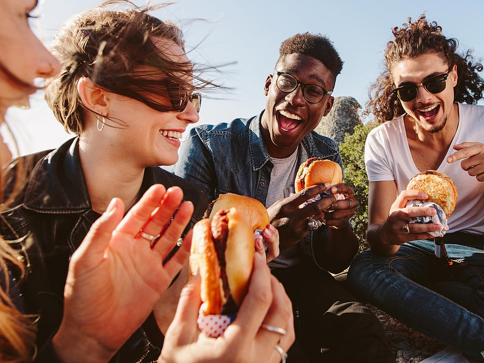 a group of young adults sitting in a group laughing, smiling and eating burgers