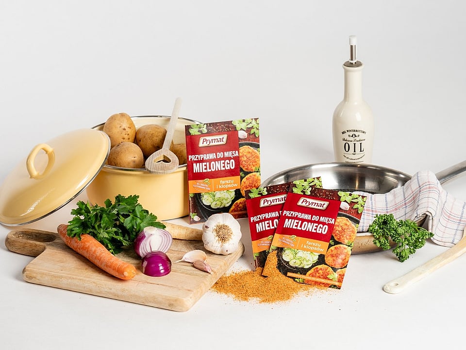 Assorted cooking ingredients arranged on a white background, including potatoes in a yellow pot, fresh vegetables like carrots, parsley, onions, and garlic on a wooden board, packets of Prymat seasoning for minced meat, a frying pan with a kitchen towel, and an olive‑oil bottle.