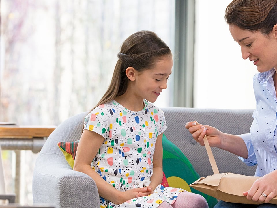 A child and an adult sit together on a sofa in a bright living room. The adult is opening a cardboard package, while the child watches attentively. Soft furnishings and natural light create a warm, home‑like atmosphere.