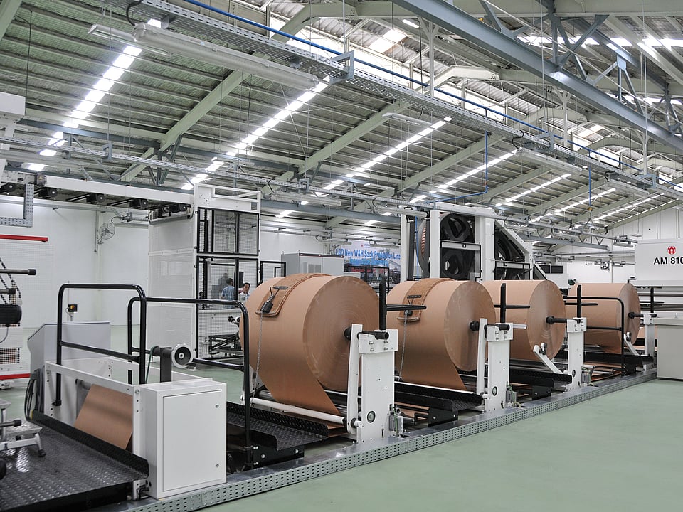 Industrial production line with large rolls of brown paper mounted on automated unwinding machines inside a factory hall.