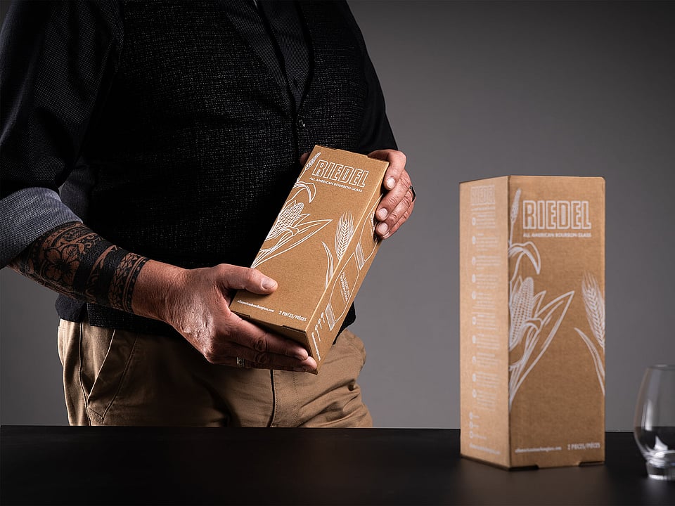 man in black clothing holding a brown corrugated box with a glass on the table next to him
