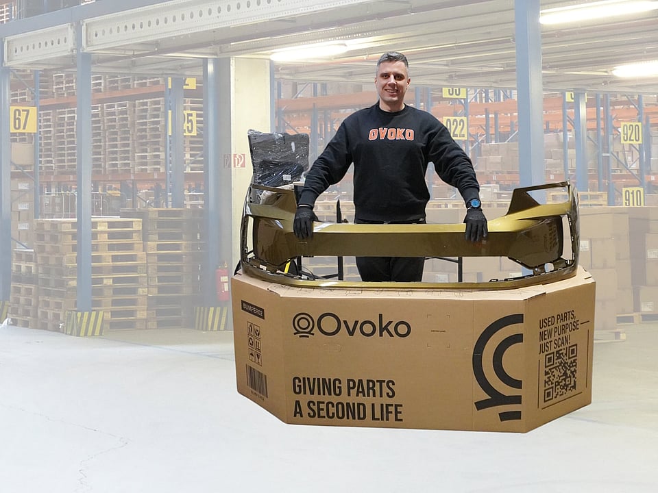 Person standing behind a large cardboard shipping box while holding a gold-coloured automotive bumper part placed across the top. The box is printed with the “Ovoko” logo and the text “Giving parts a second life,” along with a QR code and handling icons. The person is wearing dark clothing and gloves. Additional packaging material and a wrapped item are visible behind the box, all set against a plain white background.
