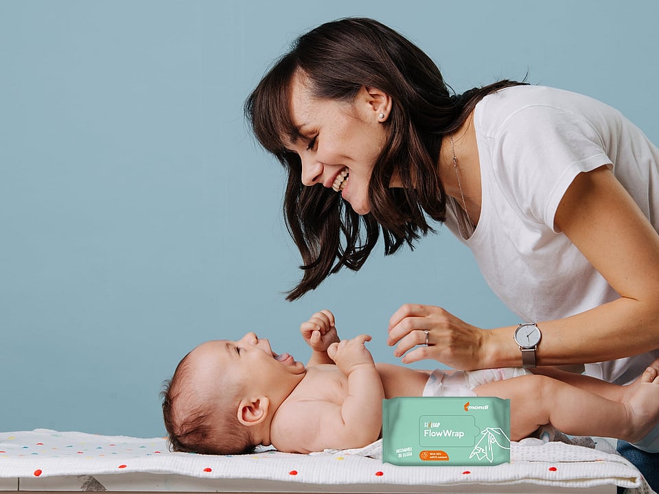 Person leaning over a baby lying on a changing mat in an indoor setting. A pack of baby wipes is visible in the foreground. The scene focuses on a caregiving moment between the person and baby, with a light blue background and soft natural lighting.