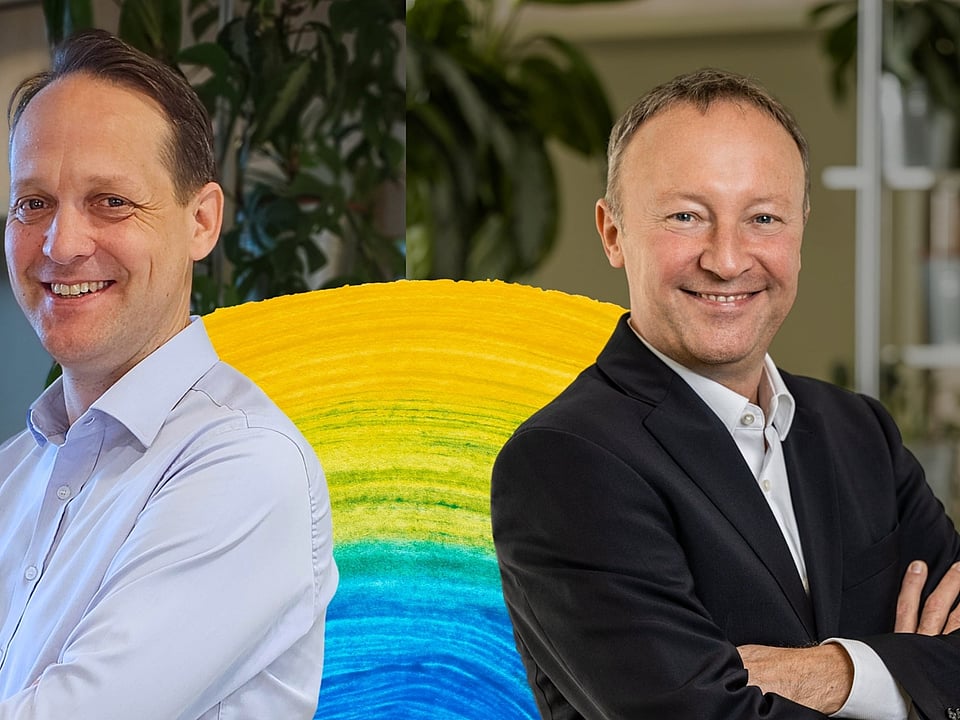 Two people standing with folded arms in front of a colourful circular display, with indoor plants and office elements visible in the background.