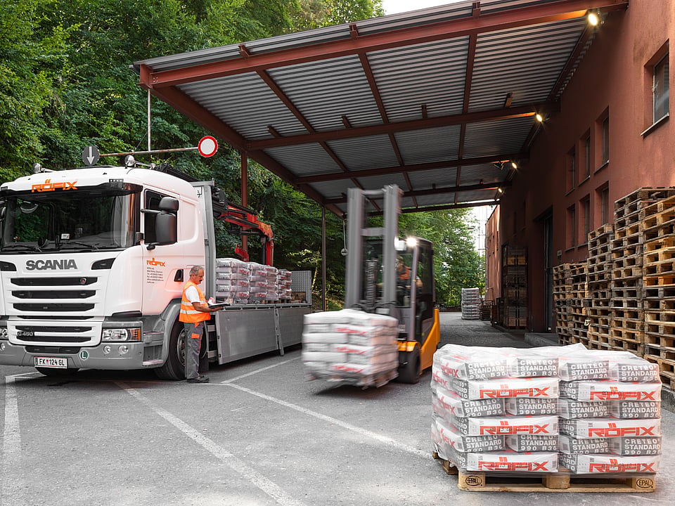 Delivery truck parked under a covered loading area while a forklift transports wrapped pallets. Stacks of palletised building materials are arranged in the foreground, with additional wooden pallets stored beside an industrial building surrounded by trees.