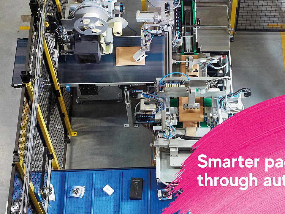 Overhead view of an automated packaging production line in an industrial facility, with machinery processing cardboard packages. A pink graphic overlay in the foreground displays the text: “Smarter packaging through automation.”