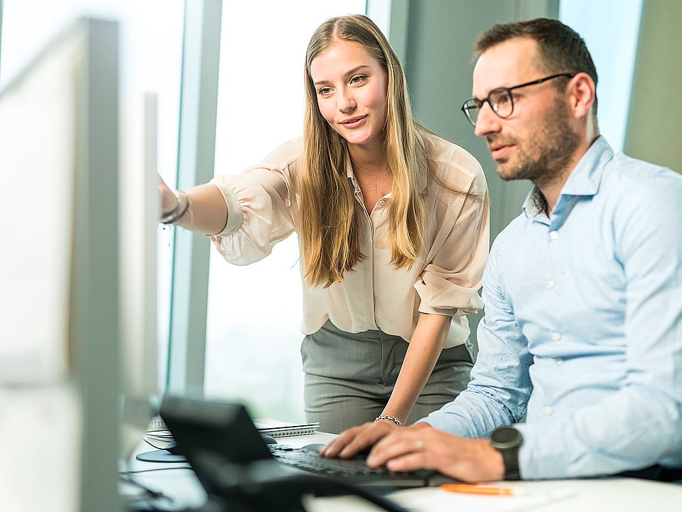 A man and a woman look closely at a computer screen; the woman points at it while both appear highly focused and engaged.