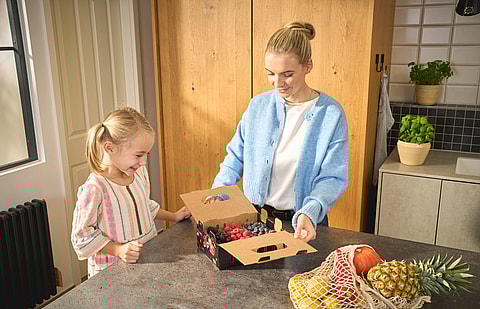 A woman and a child stand next to a kitchen island, opening a wrap-around container for fruits made of corrugated board.