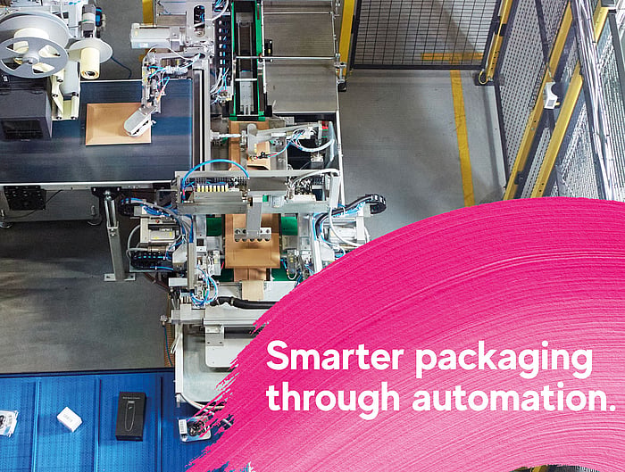 Overhead view of an automated packaging production line in an industrial facility, with machinery processing cardboard packages. A pink graphic overlay in the foreground displays the text: “Smarter packaging through automation.”