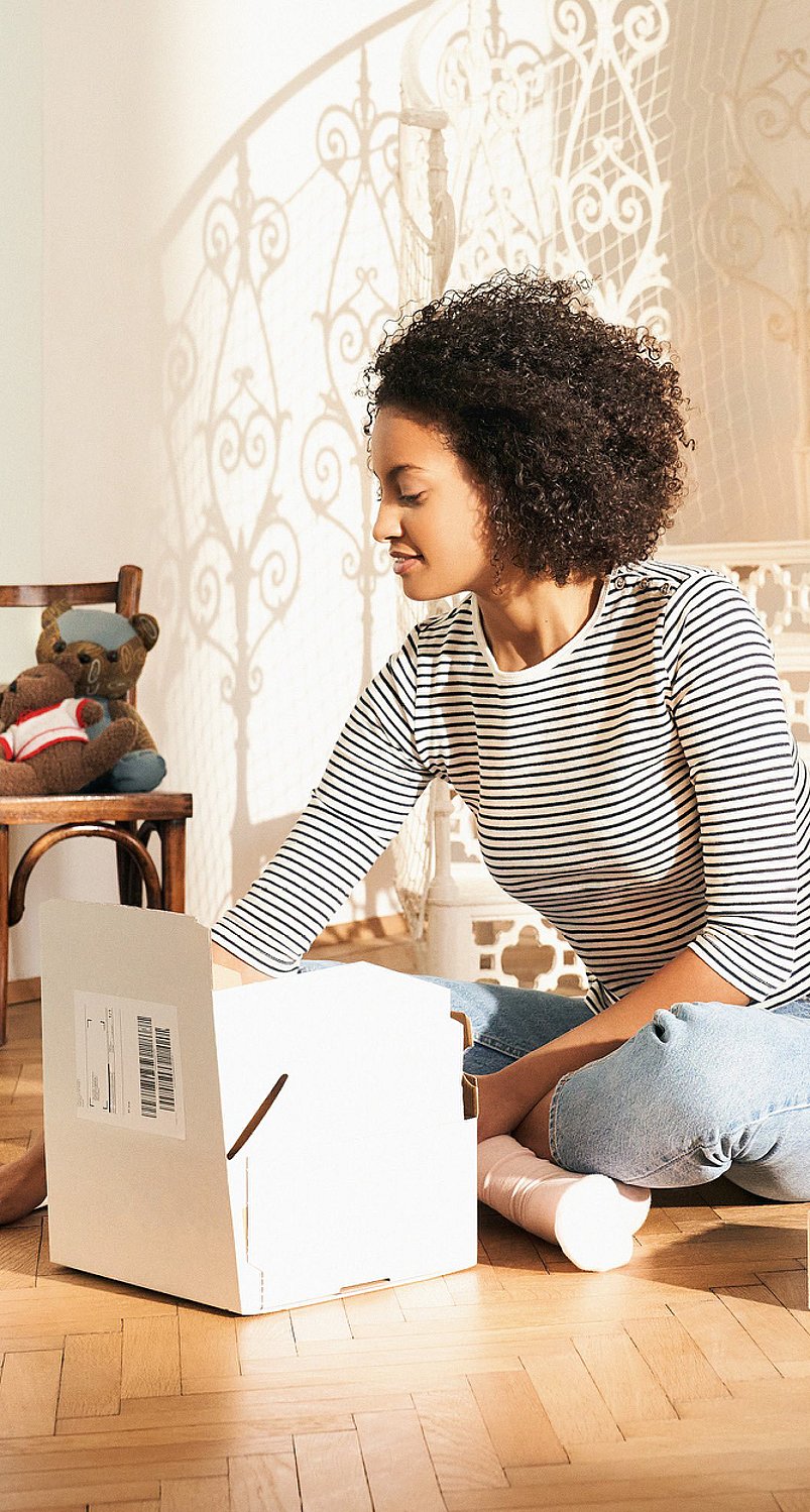 A woman sitting on the floor opening a corrugated box. Other corrugated boxes surround her, as well as a paper mailer bag and filling material made of paper.