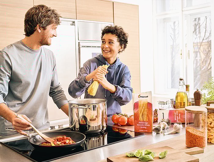Erwachsene Person hält eine Einkaufstasche aus Papier mit Verpackungssymbolen neben Lebensmitteln auf einer Küchenarbeitsfläche.