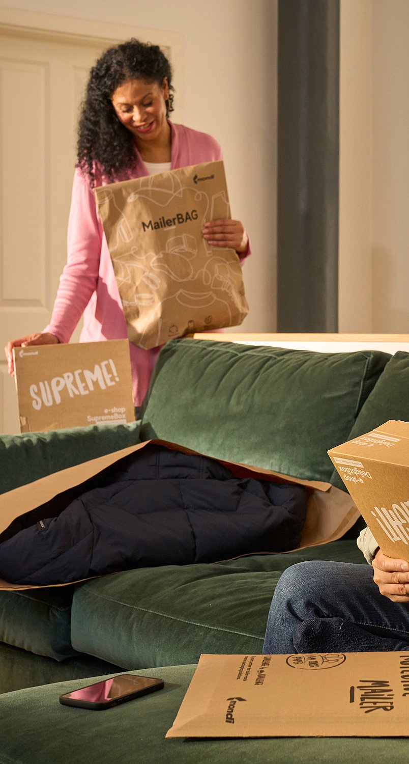 Ein Mann sitzt auf dem Sofa und packt eine Mondi DelightBox mit einem Protective Mailer vor sich aus, während eine Frau mit einem MailerBag und einer SupremeBox in der Hand im Hintergrund steht.