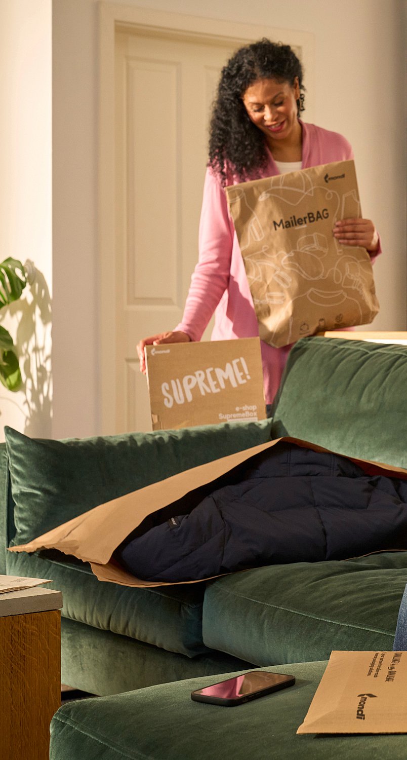 A man is sitting on the sofa unpacking a Mondi DelightBox with a Protective Mailer in front of him, while a woman stands in the back holding a MailerBag and a SupremeBox.
