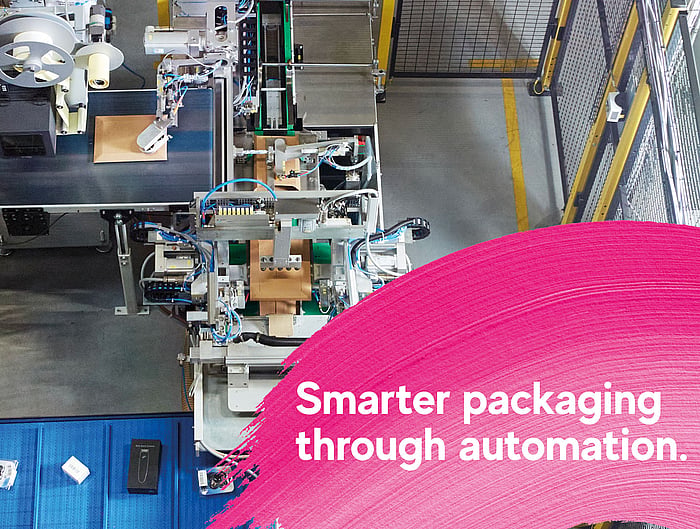 Aufsicht auf eine automatisierte Verpackungsproduktionslinie in einer Industrieanlage, auf der Maschinen Kartonverpackungen verarbeiten. Im Vordergrund zeigt eine pinke grafische Einblendung den Text: „Smarter packaging through automation.“