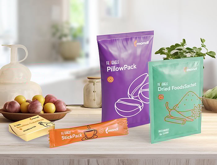 Various colorful packaging samples are displayed on a wooden kitchen-style countertop, including a purple pillow pack, a green dried food sachet, an orange stick pack, and two small butter-like wrapped items. Fresh fruit and a potted plant sit in the background.