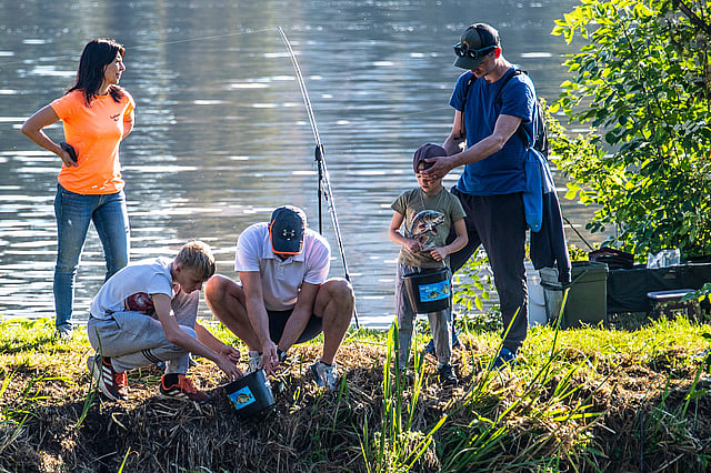 A group of people fishing together beside a lakeshore; two adults help a child prepare fishing gear while another child watches, and a woman stands nearby enjoying the scene.