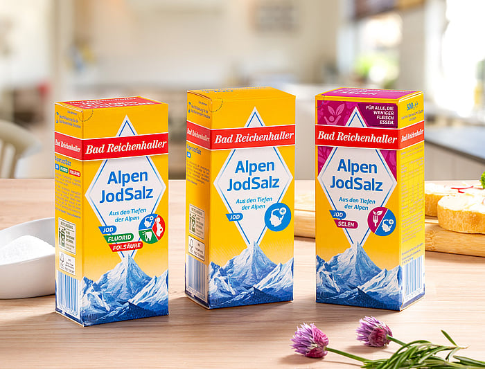 Three cartons of “Alpen JodSalz” iodized salt stand on a wooden kitchen counter. The packages feature bright yellow, red, and blue designs with an illustration of alpine mountains. Fresh herbs and kitchen items are softly blurred in the background.
