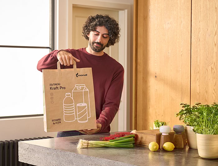 Adult holding a paper shopping bag with packaging symbols, standing next to groceries on a kitchen counter.
