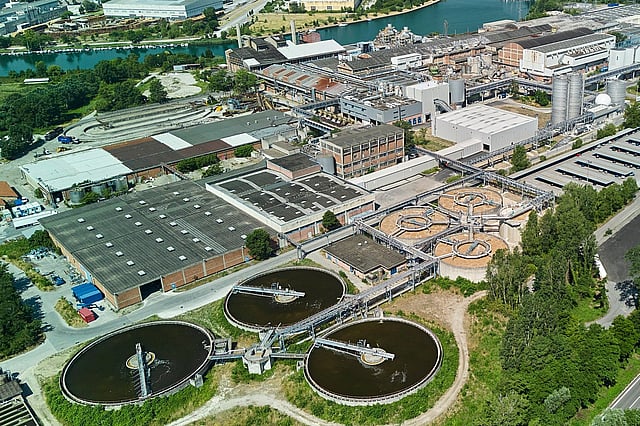 Aerial view of an industrial facility with large circular water-treatment basins, warehouses, and surrounding greenery near a river.