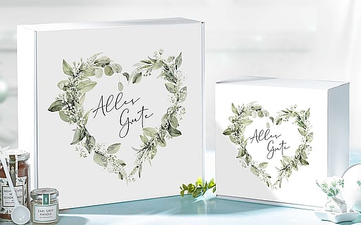 A white corrugated gift box featuring a floral heart-shaped design and the inscription in German,