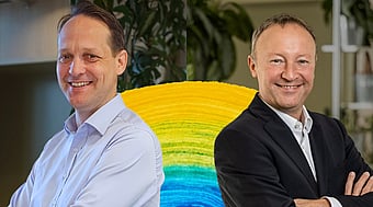 Zwei Personen mit verschränkten Armen stehen vor einer farbenfrohen kreisförmigen Installation. Im Hintergrund sind Zimmerpflanzen und Elemente einer Büroumgebung zu sehen.