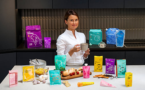 Person standing behind a table displaying packaged food products and holding a mug. Multiple colourful product packages are arranged on shelves and across the table alongside snacks and ingredients in a modern indoor setting.