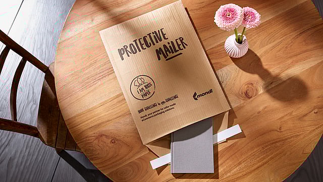 A Protective Mailer is open on a table, with a book half-packed into it and a vase with a flower next to it.