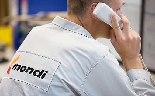 A person wearing a light grey Mondi work shirt speaks on a corded phone inside an office or industrial workspace.