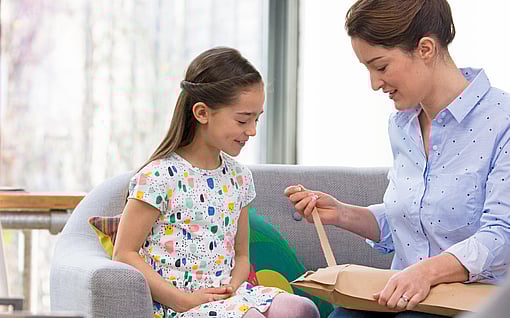 A child and an adult sit together on a sofa in a bright living room. The adult is opening a cardboard package, while the child watches attentively. Soft furnishings and natural light create a warm, home‑like atmosphere.