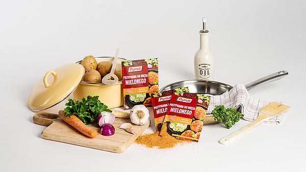 Assorted cooking ingredients arranged on a white background, including potatoes in a yellow pot, fresh vegetables like carrots, parsley, onions, and garlic on a wooden board, packets of Prymat seasoning for minced meat, a frying pan with a kitchen towel, and an olive‑oil bottle.
