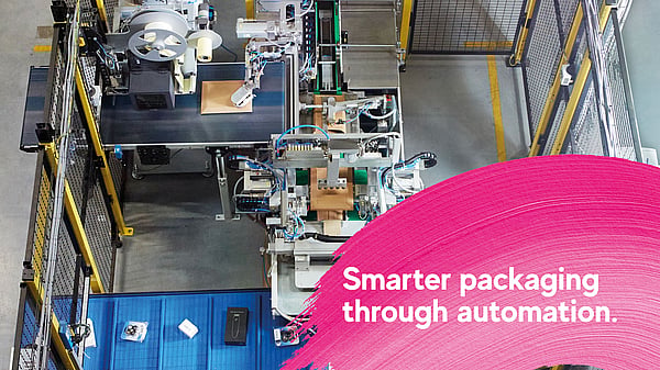 Overhead view of an automated packaging production line in an industrial facility, with machinery processing cardboard packages. A pink graphic overlay in the foreground displays the text: “Smarter packaging through automation.”