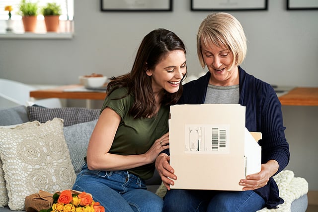 Two people sitting together on a sofa, opening and looking at a cardboard delivery box with a shipping label. The scene takes place in a home setting and highlights paper-based packaging used for parcel delivery.