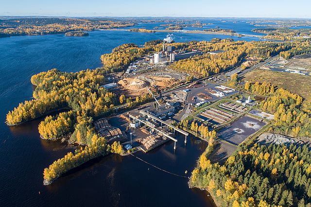 Aerial view of an industrial facility surrounded by autumn‑coloured forests and waterways, with processing buildings, storage areas, and access roads spread across the landscape.
