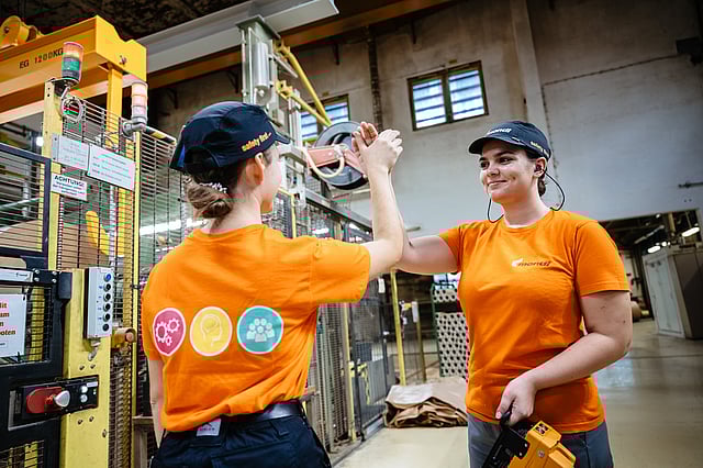 Two Mondi workers wearing orange shirts and caps stand in an industrial facility, giving each other a high‑five near machinery and safety equipment.